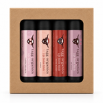 A boxed set of four Tagi Organics lip balm sticks in a kraft paper gift box. The lip balms come in four shades: candy hearts in pink, XOXO in peach, lip balm stick in red, and lip balm stick in mauve.