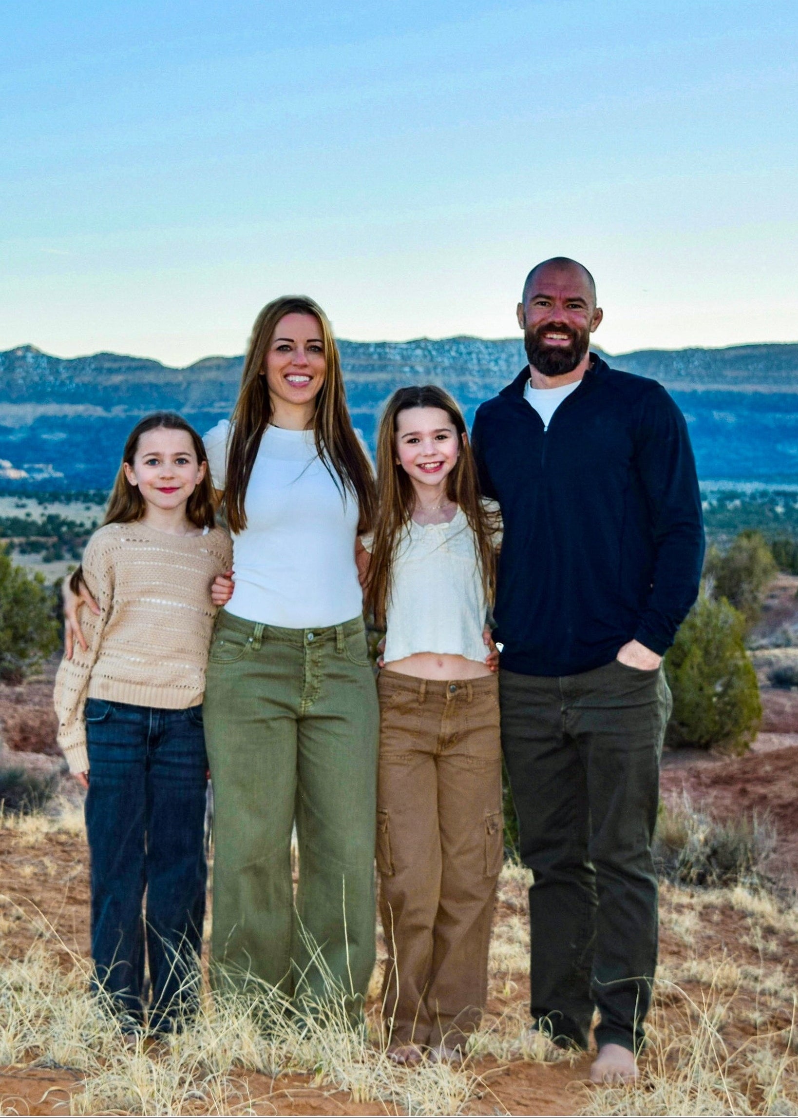 A family of four stands together outdoors on a dry, reddish-brown hillside with a vast desert landscape and mountains in the background under a clear blue sky.