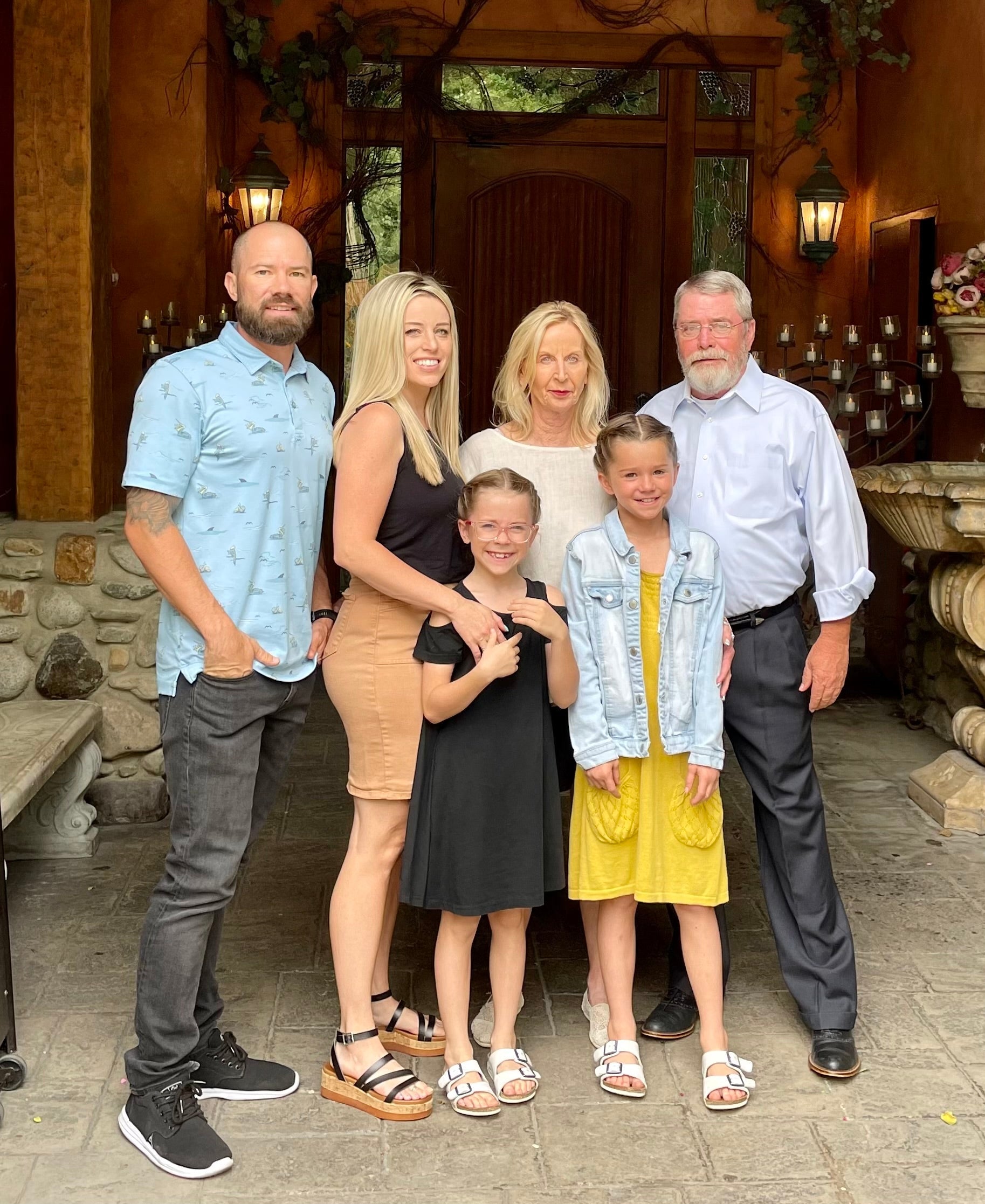 A family of six posing together in front of a rustic stone and wood entryway. The group includes two adults and four children wearing casual summer clothing, with one child in a yellow dress and another in a black dress.