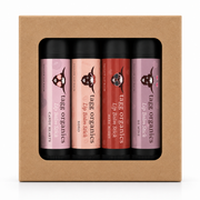 A boxed set of four Tagi Organics lip balm sticks in a kraft paper gift box. The lip balms come in four shades: candy hearts in pink, XOXO in peach, lip balm stick in red, and lip balm stick in mauve.