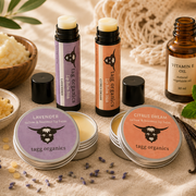 A styled flat-lay of Tagg Organics lip care products on a beige fabric background with natural props. Visible items include two lip balm tubes, two round lip balm tins labeled Lavender and Citrus Dream, a small amber bottle labeled Vitamin E Oil, and bowls of cream and botanical ingredients.