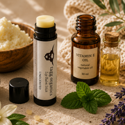 A skincare set featuring a 'Taze Organics' natural body butter stick, a Vitamin E oil bottle, and a small glass bottle, styled with vanilla beans, lavender, mint, and bowls of ingredients on a beige knit blanket.
