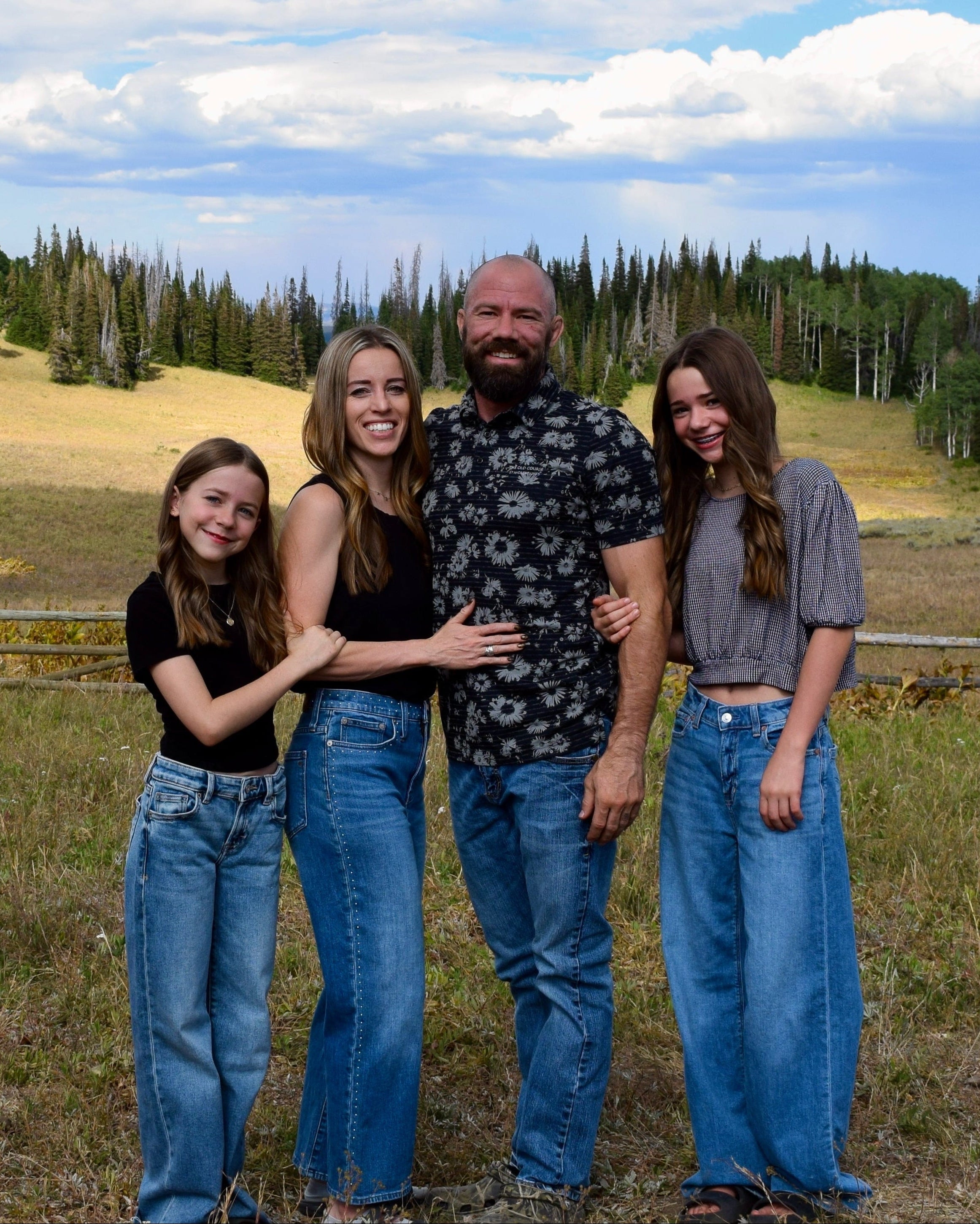 A family of four posing together outdoors in a grassy field with hills, trees, and a blue sky with clouds in the background.