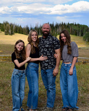 A family of four posing together outdoors in a grassy field with hills, trees, and a blue sky with clouds in the background.