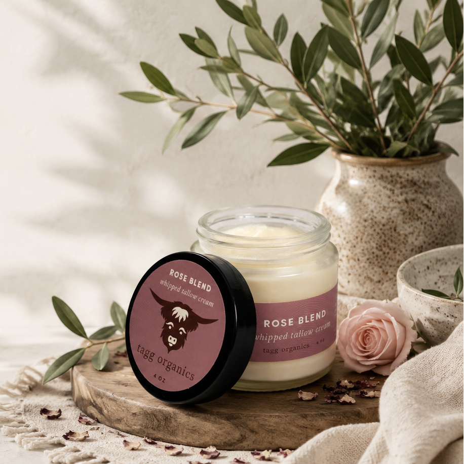 A candle jar labeled 'Rose Blend' with its lid placed beside it on a wooden tray, styled with olive branches, a pink rose, and soft natural shadows on a light background.